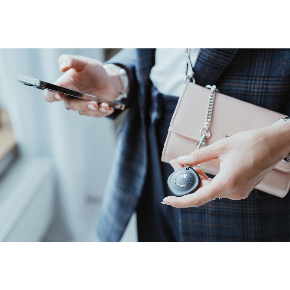 Person hält schwarzen, runden Verbatim Bluetooth Tracker und ein Smartphone. Eine rosafarbene Handtasche hängt über der.