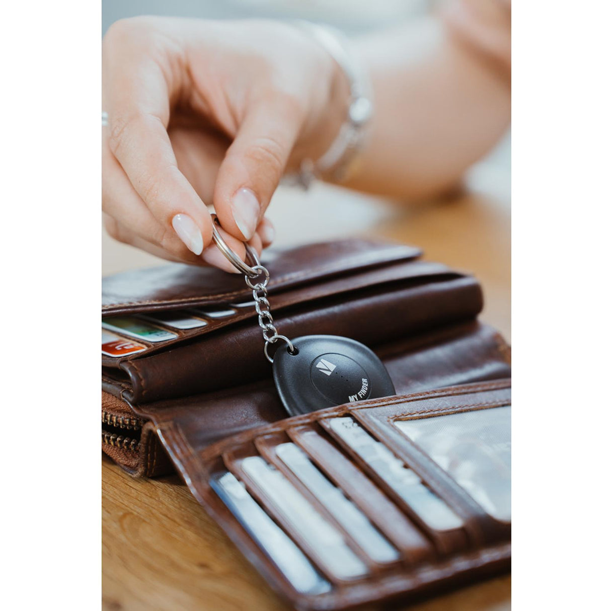 Hand hält schwarzen Verbatim Bluetooth Tracker am Schlüsselring über einer braunen Ledergeldbörse mit Kreditkarten.