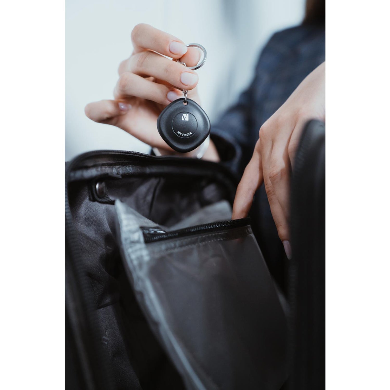 Frauenhand hält schwarzen, runden Verbatim Bluetooth Tracker mit Schlüsselring über offener, schwarzer Tasche.