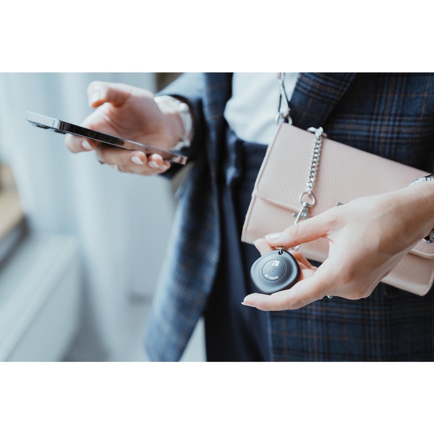 Nahaufnahme: Eine Person hält einen schwarzen, runden Bluetooth-Tracker und ein Smartphone. Eine rosa Handtasche ist.