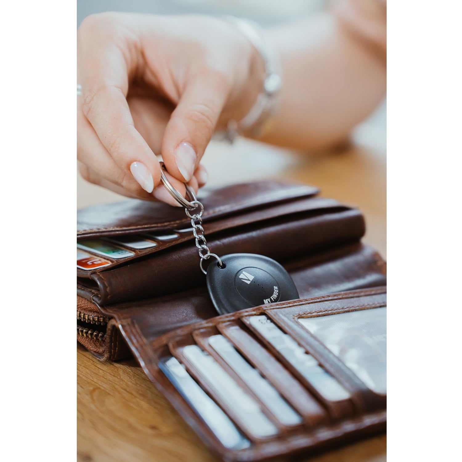 Hand hält schwarzen, runden Verbatim Bluetooth Tracker mit silbernem Schlüsselring über braunem Lederportemonnaie.