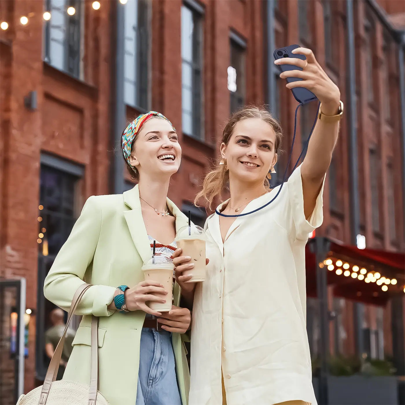 Zwei Frauen mit Kaffee, eine macht Selfie mit blauem iPhone 16 mit Kette.
