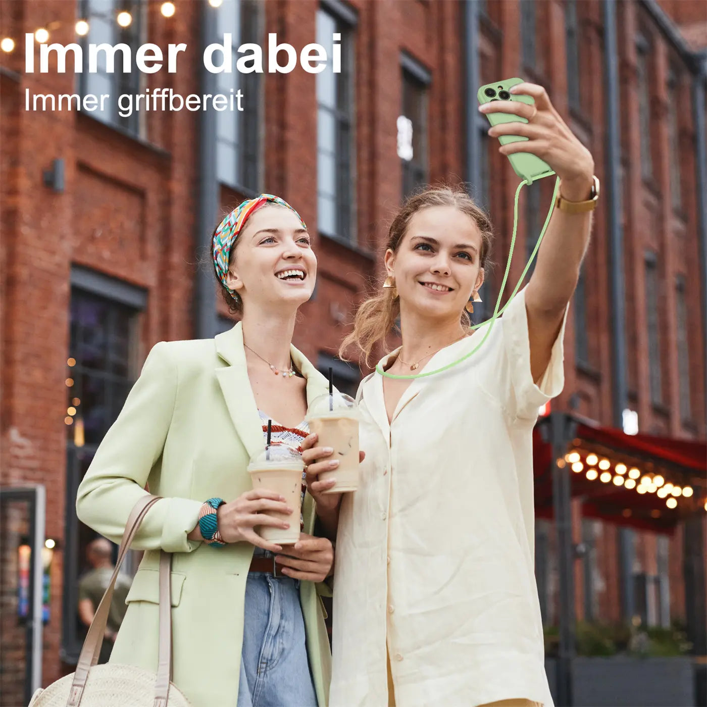 Zwei Frauen mit Kaffee, eine macht Selfie mit hellgrünem Handy mit Kette.
