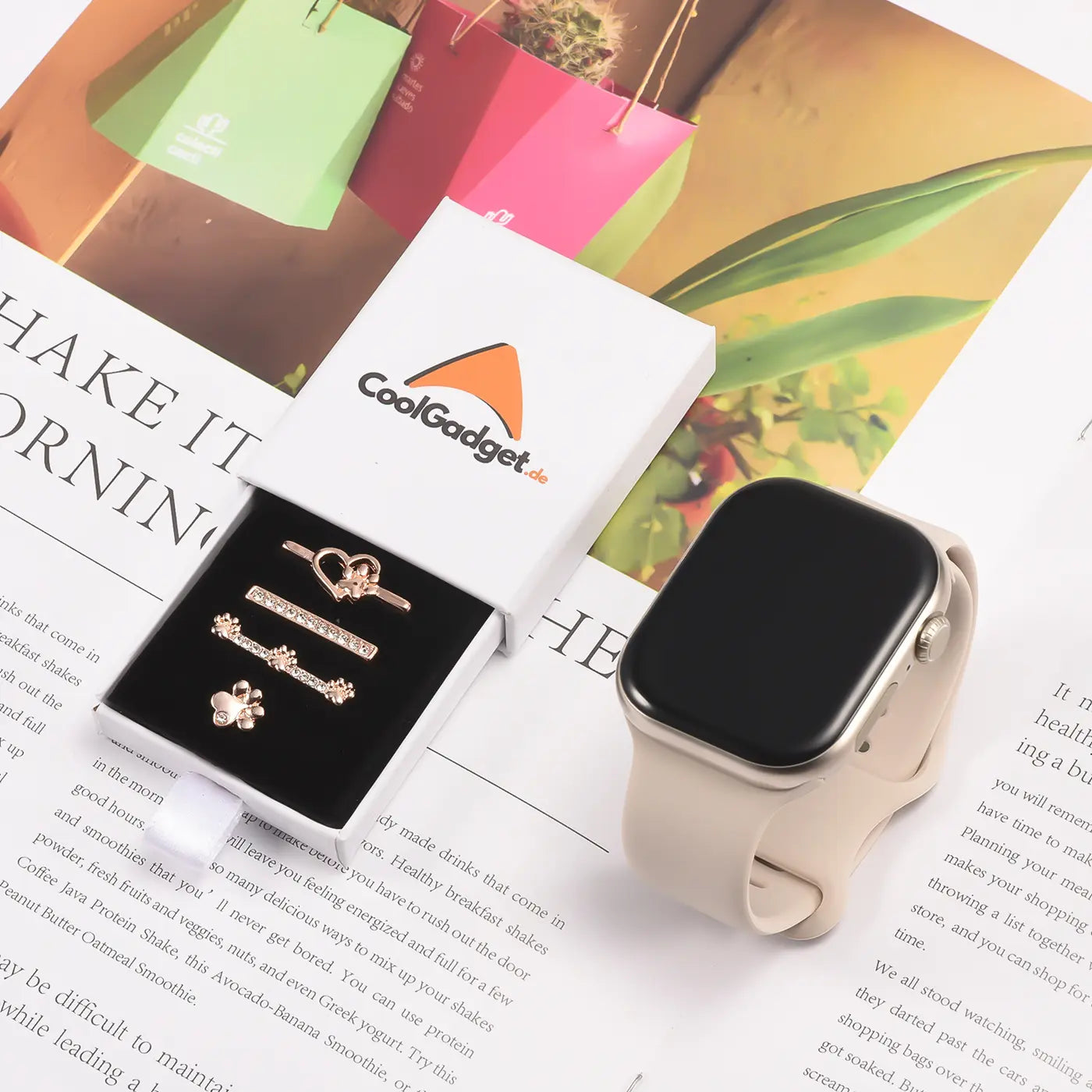 Goldfarbene Smartwatch-Armbandschmuck-Charms in Schachtel, neben einer hellbeigen Smartwatch.