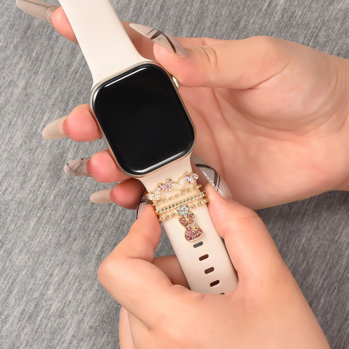 Hände mit Smartwatch in Beige, Armband mit goldenen Charms (Krönchen, Teddybär) und rosa Steinen.