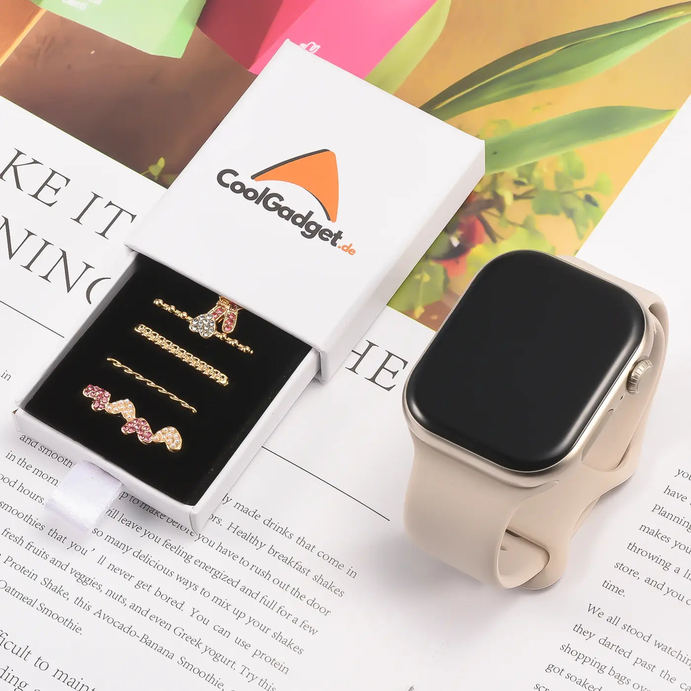 Goldfarbener Smartwatch-Schmuck mit rosa/roten Steinen in Box, daneben Smartwatch.