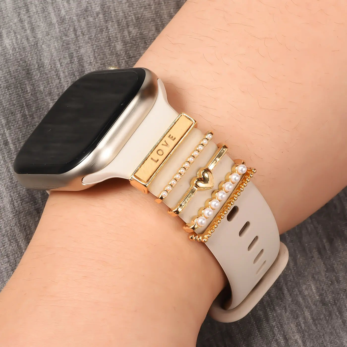 Smartwatch-Armband mit goldfarbenen Charms (Herz, Perlen, "LOVE") an beigefarbenem Silikonband, Nahaufnahme.