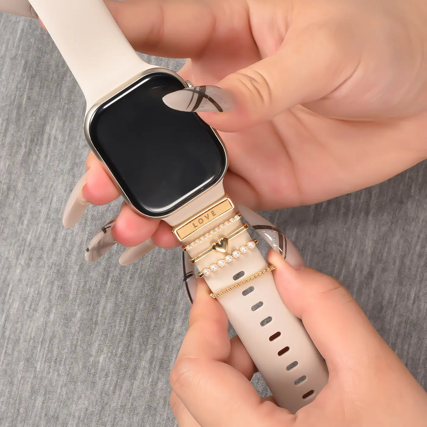 Hände befestigen goldene Charms an einem hellbeigen Smartwatch-Armband.