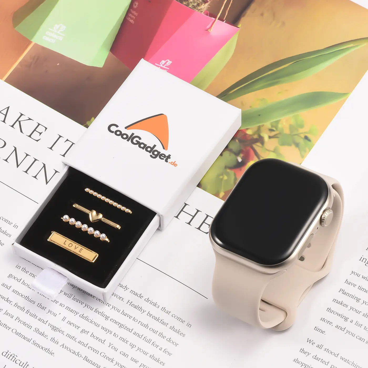 Goldene Smartwatch-Charms mit "LOVE"-Schriftzug in Box neben beiger Smartwatch.