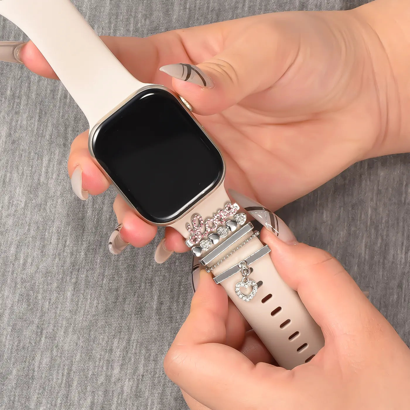 Smartwatch-Armband (beige) mit silbernen Charms (Herz, "Love") und rosa Steinen, Nahaufnahme.