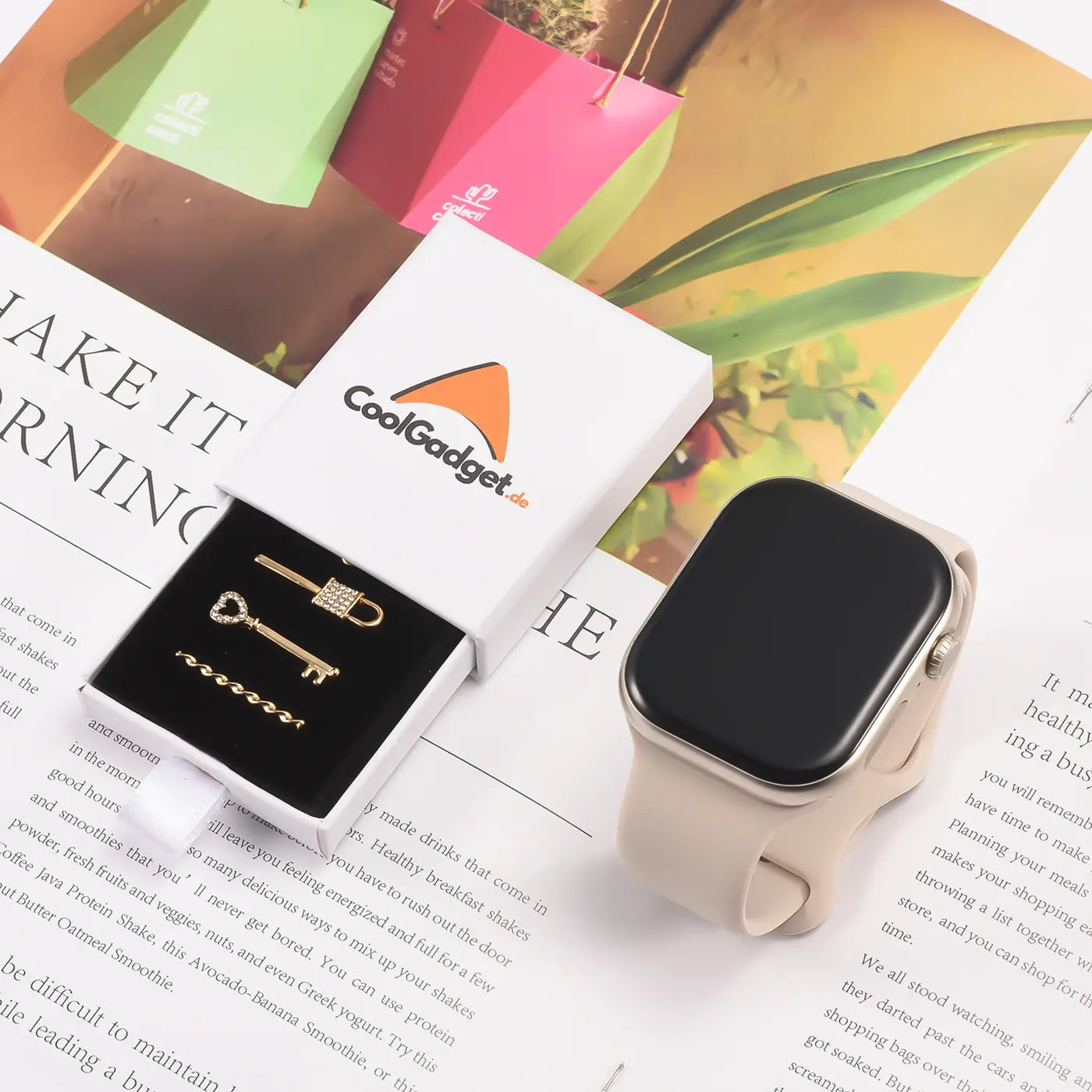 Goldfarbene Smartwatch-Charms (Schlüssel, Schloss) in weißer Box neben beiger Smartwatch, Draufsicht.