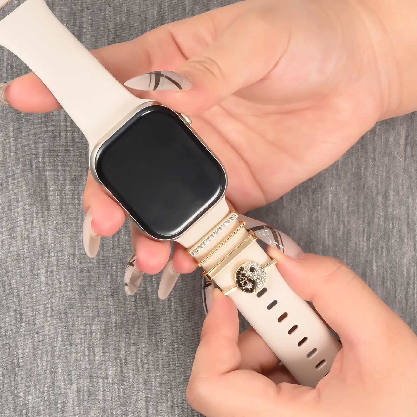 Hände mit hellbeigem Smartwatch-Armband, goldfarbenen Charms und Kristallen, Nahaufnahme.