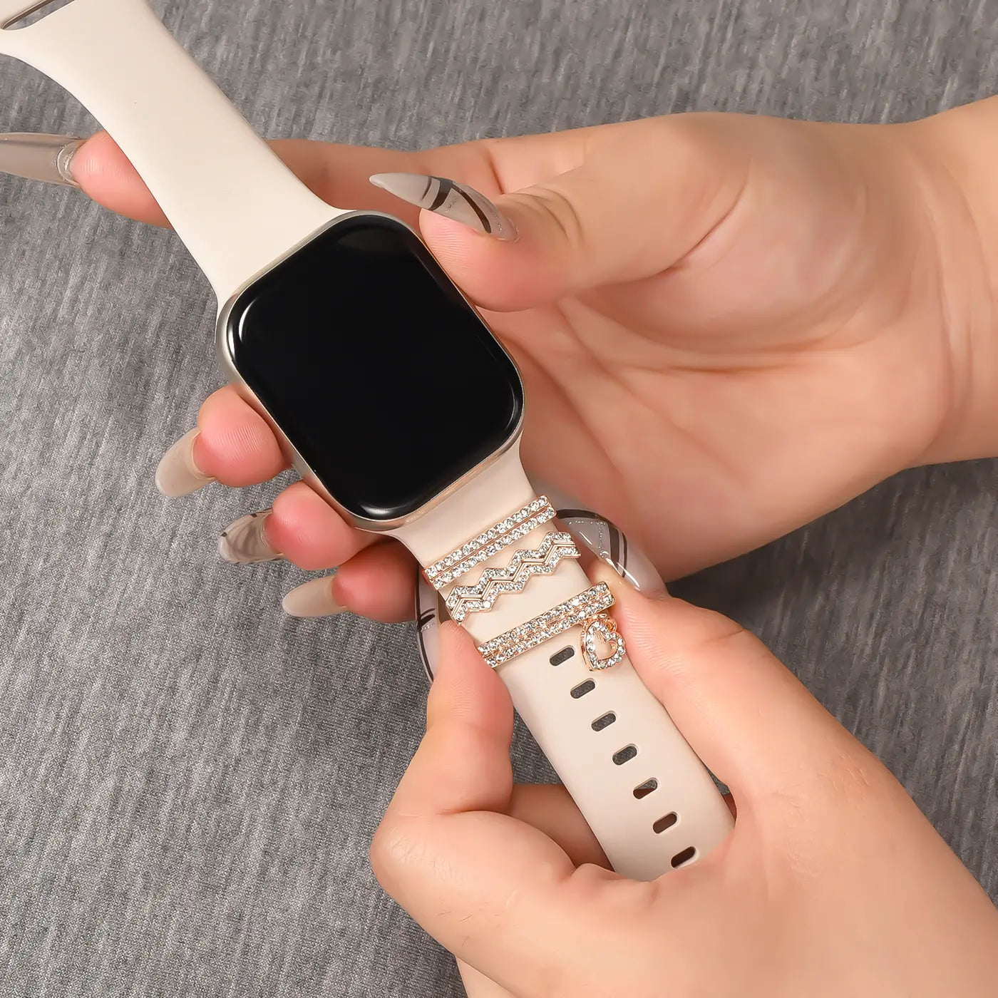 Hände befestigen silbernen Schmuck mit Strasssteinen an einem hellbeigen Smartwatch-Armband.