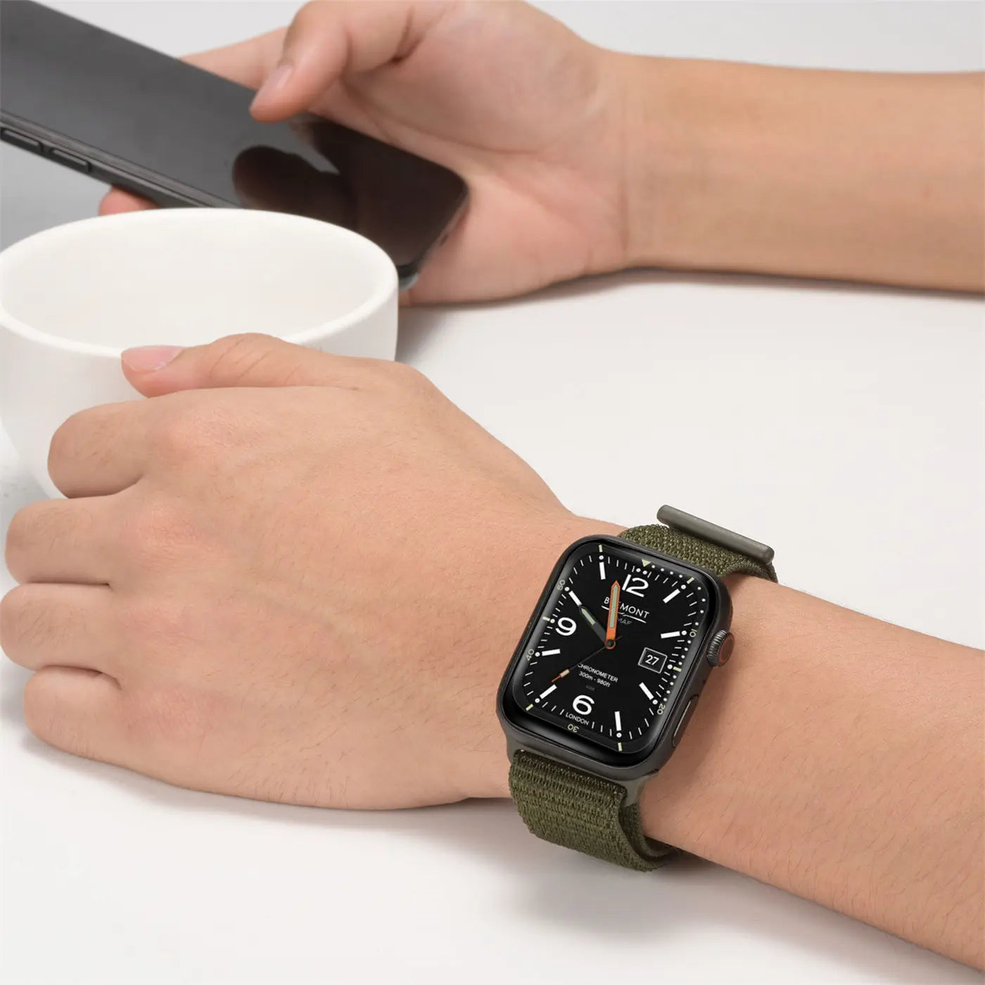 Nahaufnahme: Handgelenk mit schwarzer Apple Watch und olivgrünem Nylon-Armband, Handy und Tasse im Hintergrund.