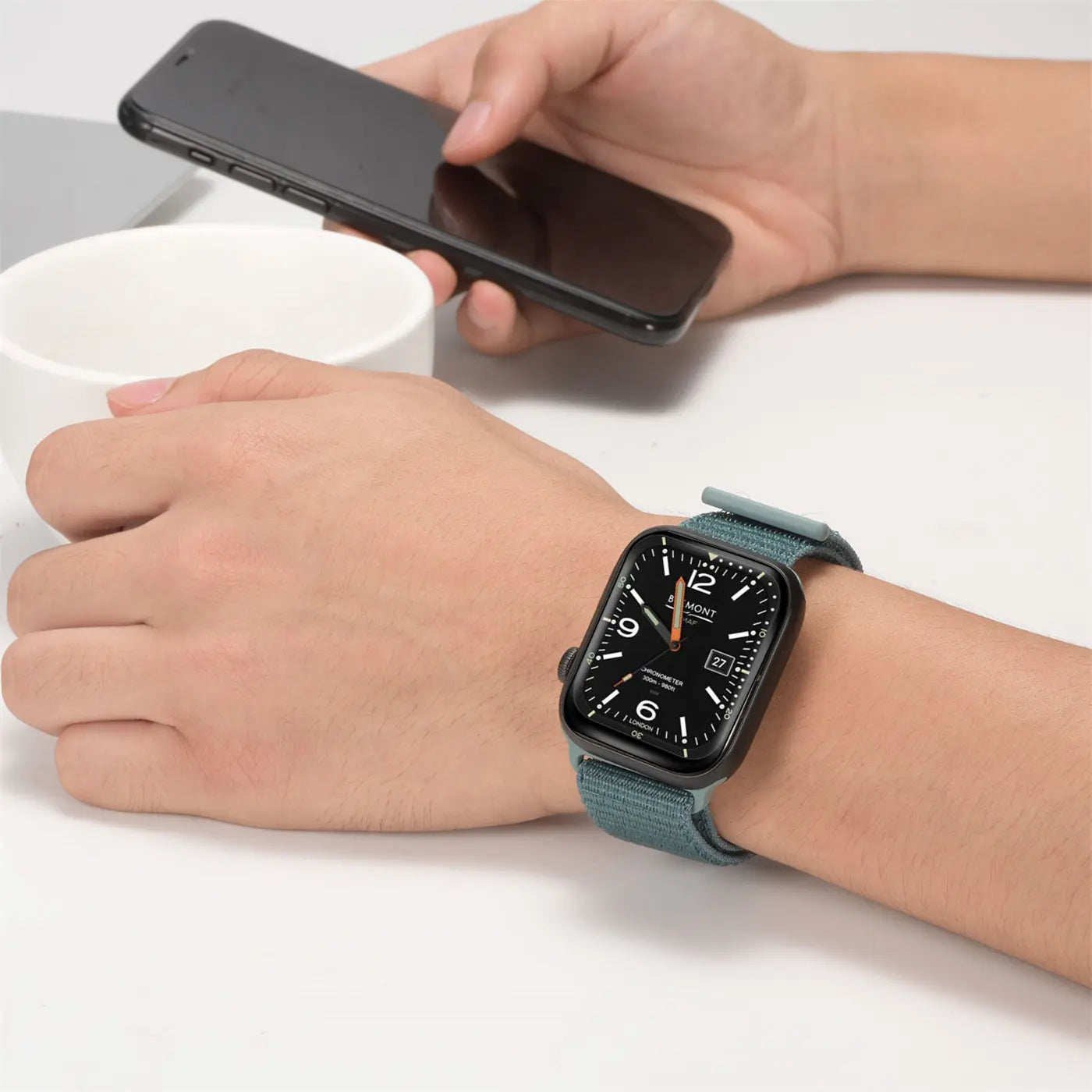 Handgelenk mit Apple Watch in Schwarz und blaugrünem Nylon-Armband, daneben ein Smartphone.