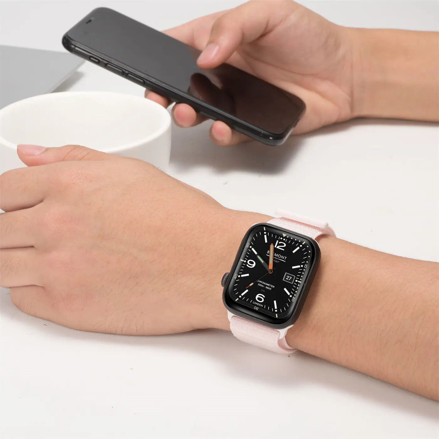 Nahaufnahme: Handgelenk mit schwarzer Apple Watch und hellrosa Nylon-Armband. Im Hintergrund ein Smartphone.