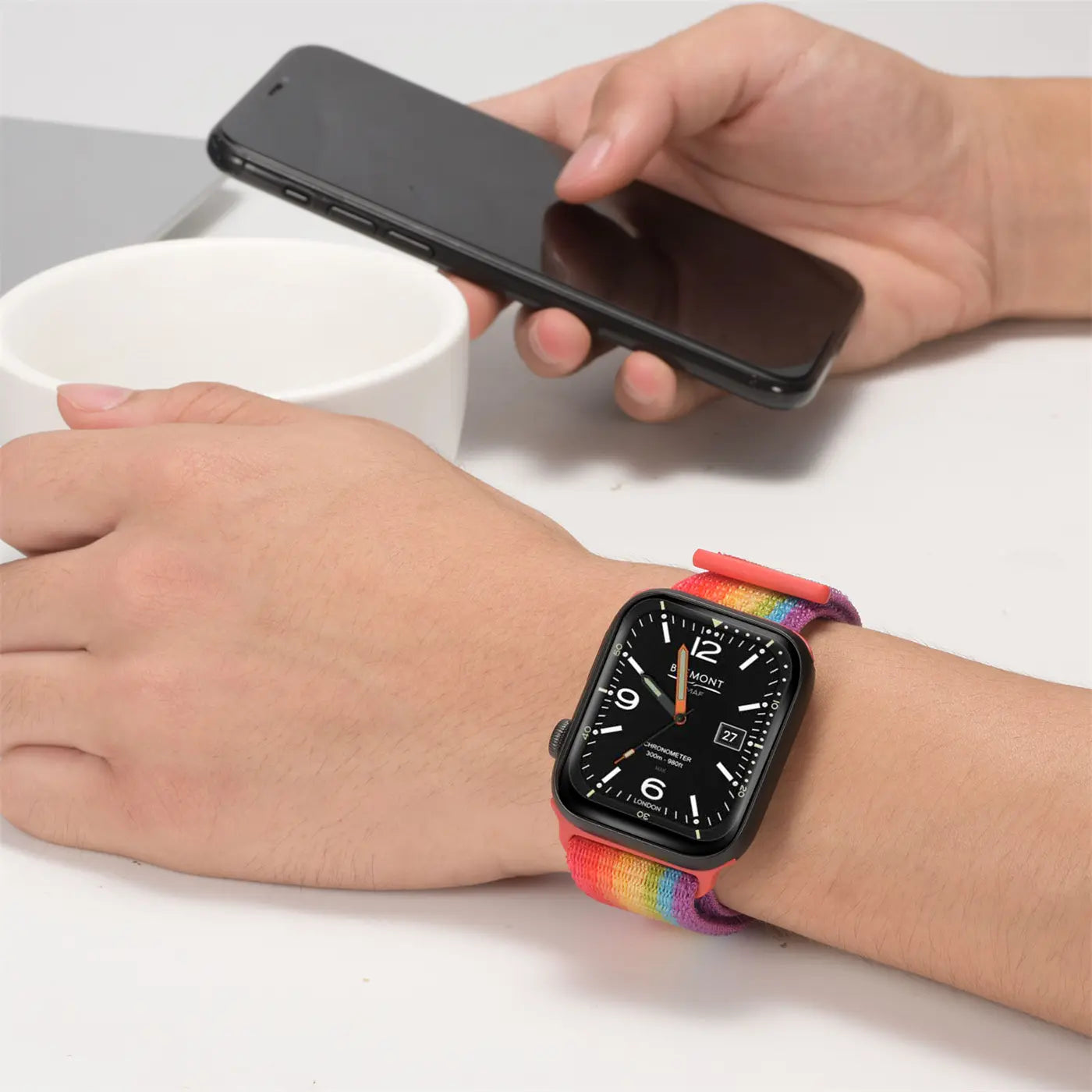 Nahaufnahme: Eine Apple Watch mit schwarzem Gehäuse und regenbogenfarbenem Nylon-Fitnessarmband am Handgelenk.