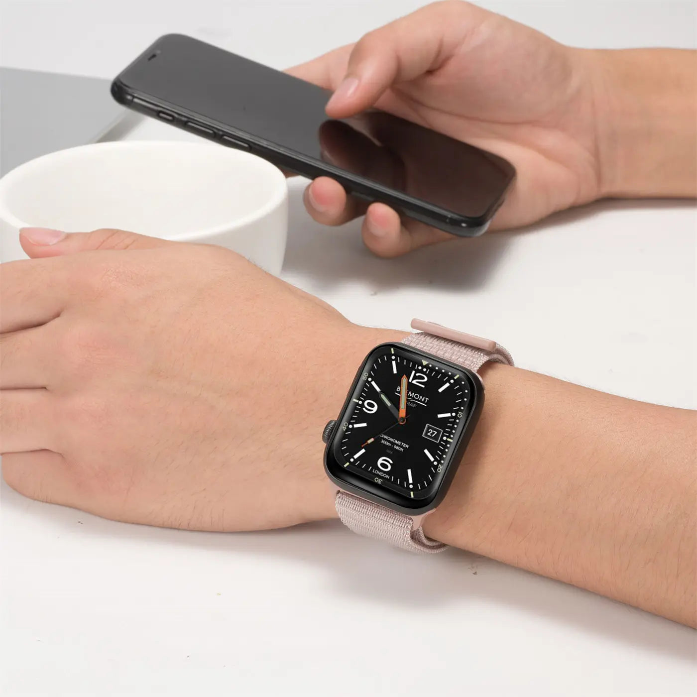Nahaufnahme: Handgelenk mit Apple Watch und rosafarbenem Nylonarmband. Im Hintergrund ein Smartphone.