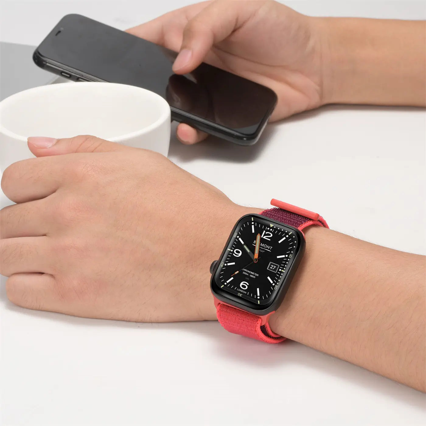 Nahaufnahme: Handgelenk mit schwarzer Apple Watch und rotem Nylon-Armband. Im Hintergrund ein Smartphone und eine Tasse.