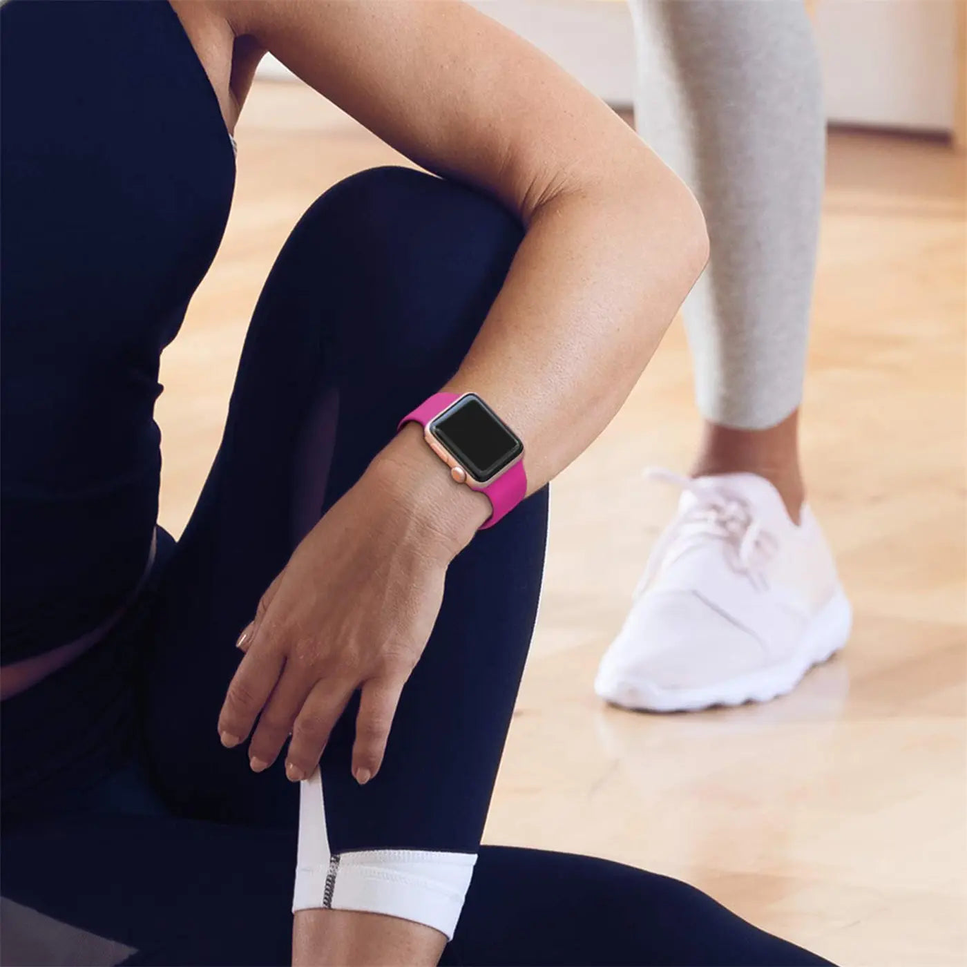 Nahaufnahme: Frau trägt pinkes TPU-Fitnessarmband mit goldener Apple Watch. Sie sitzt auf Holzboden.