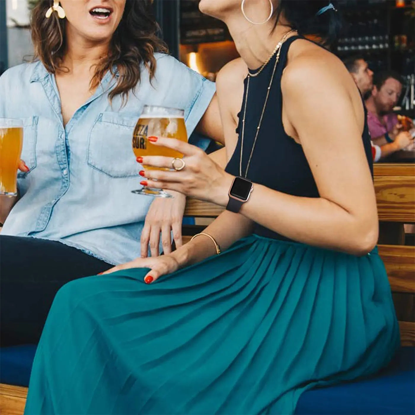 Frau mit dunkelbraunem TPU-Armband an Apple Watch am Handgelenk, hält Bierglas, sitzt seitlich.