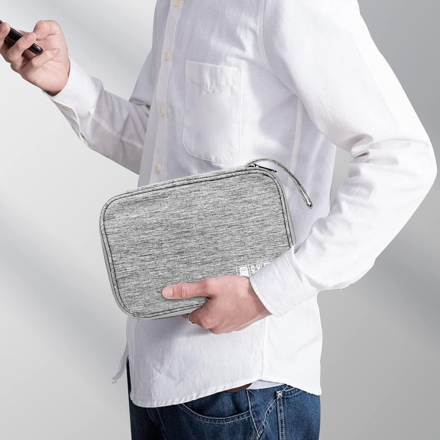 Mann hält grauen, rechteckigen Reiseorganizer aus Stoff in der Hand, ein Smartphone in der anderen. Seitenansicht.