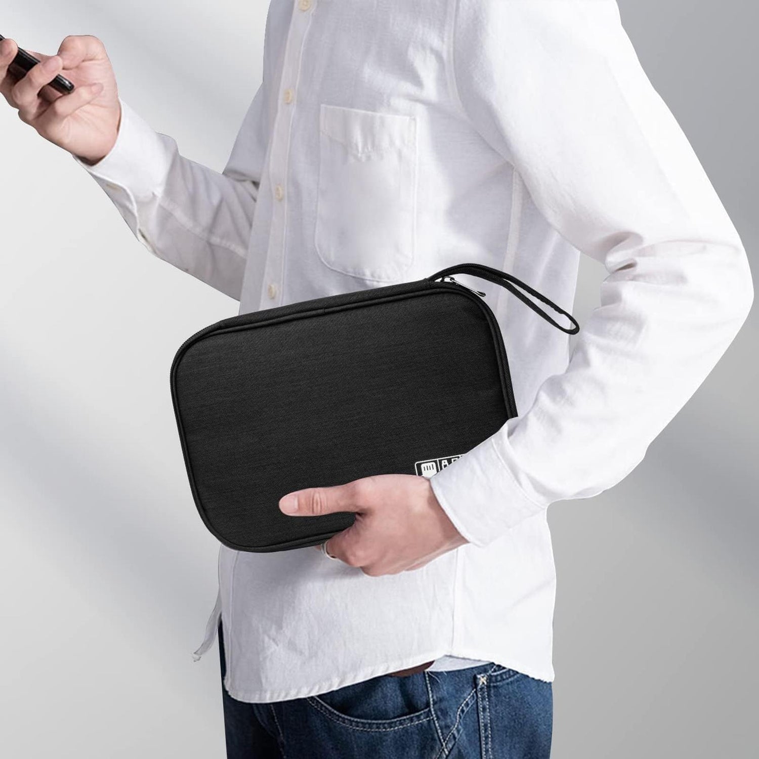 Mann hält schwarzen Reiseorganizer aus Stoff mit Handschlaufe in der linken Hand, rechts ein Smartphone. Seitenansicht.